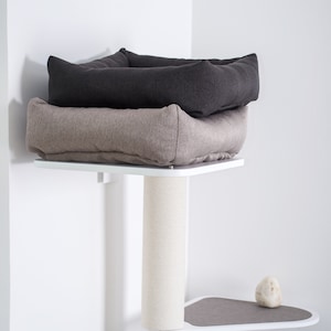 May include: A white cat tree with a beige and gray cat bed on top. The cat tree has a sisal-covered scratching post and a gray platform.