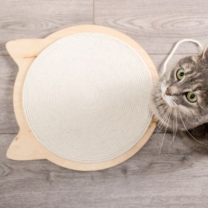 May include: A gray tabby cat looking up at a white rope-covered scratching post shaped like a cat head. The post is made of wood and has a natural finish. The cat is sitting on a light gray wood floor.