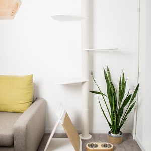 May include: White cat tree with multiple platforms and a scratching post. A grey sofa with a yellow cushion is in the background. A potted snake plant and two stainless steel dog bowls are on the floor.