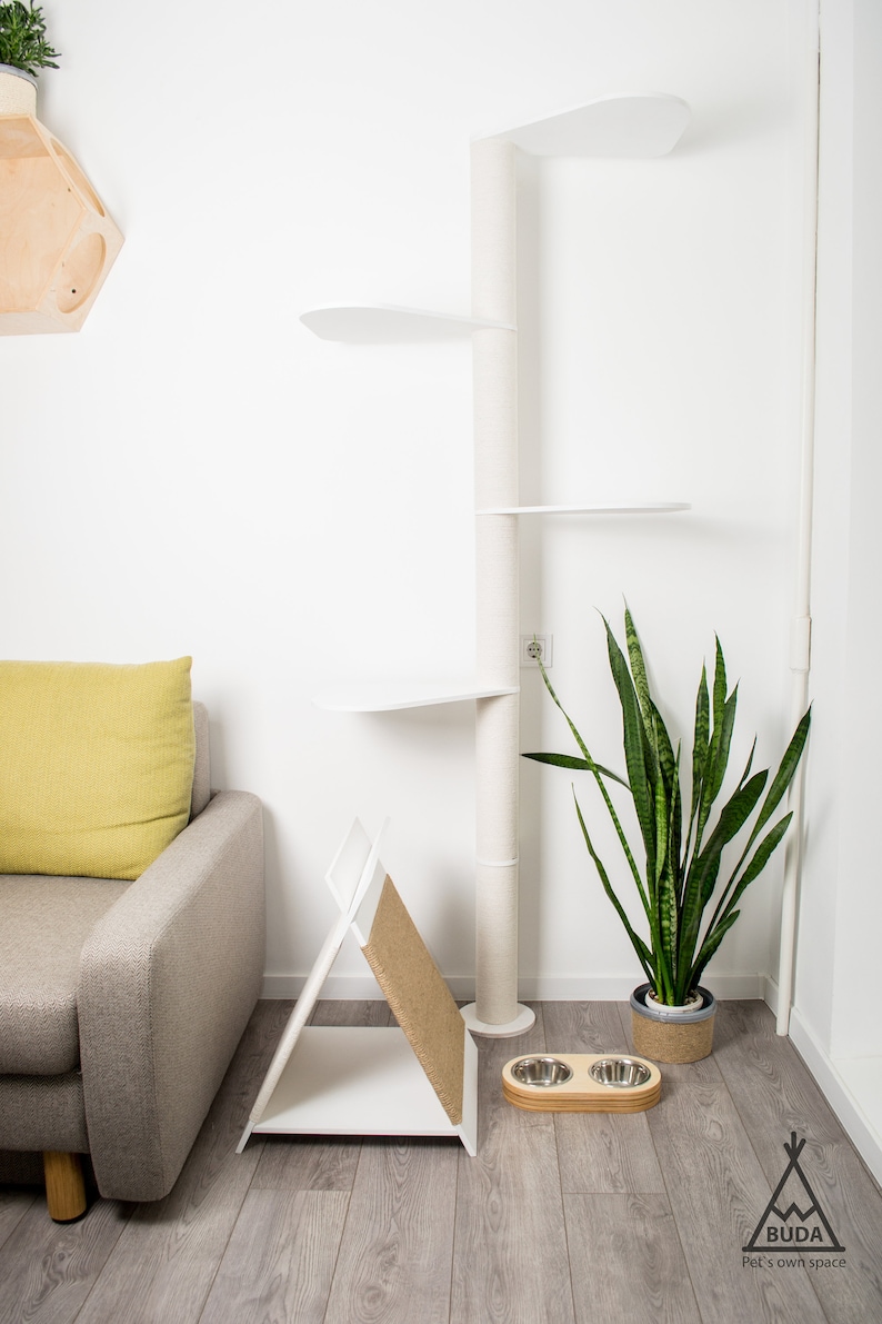 May include: White cat tree with multiple platforms and a scratching post. A grey sofa with a yellow cushion is in the background. A potted snake plant and two stainless steel dog bowls are on the floor.