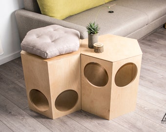 May include: Two wooden hexagonal cat furniture pieces with round openings. The piece on the left has a light gray cushion on top. The piece on the right has a small potted plant and a wooden log on top. The text "BUDA Pet's own space" is on the floor.