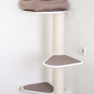 May include: White corner cat tree with three platforms and a scratching post. The platforms are covered in brown fabric. The top platform has a brown cushion. The cat tree is mounted to the wall.