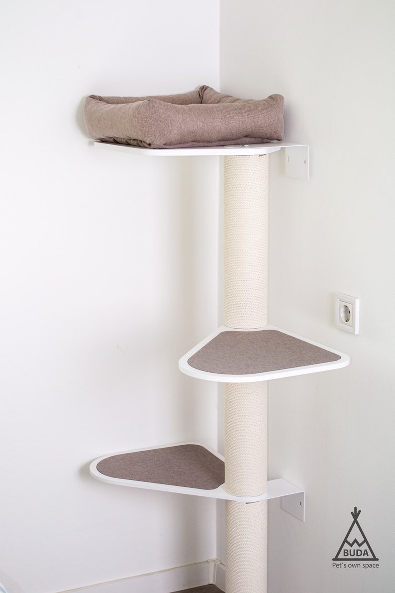 May include: White corner cat tree with three platforms and a scratching post. The platforms are covered in brown fabric. The top platform has a brown cushion. The cat tree is mounted to the wall.