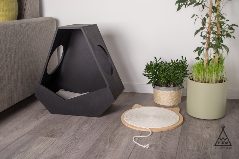 May include: A black wooden cat house with two circular openings. The house is sitting on a light wood floor. There is a white rope scratching post in front of the house.
