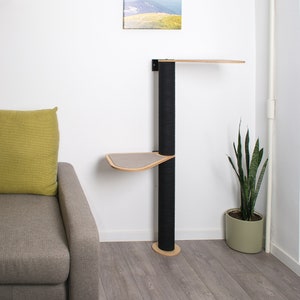 May include: A black cat tree with two platforms and a sisal-covered scratching post. The cat tree is mounted to a white wall and has a wooden base. The cat tree is in a room with a gray couch, a green pillow, and a plant in a pot.