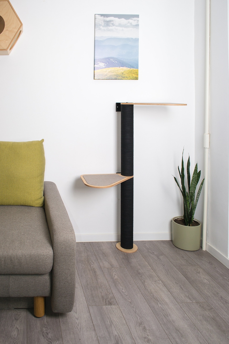 May include: A black cat tree with two platforms and a sisal-covered scratching post. The cat tree is mounted to a white wall and has a wooden base. The cat tree is in a room with a gray couch, a green pillow, and a plant in a pot.