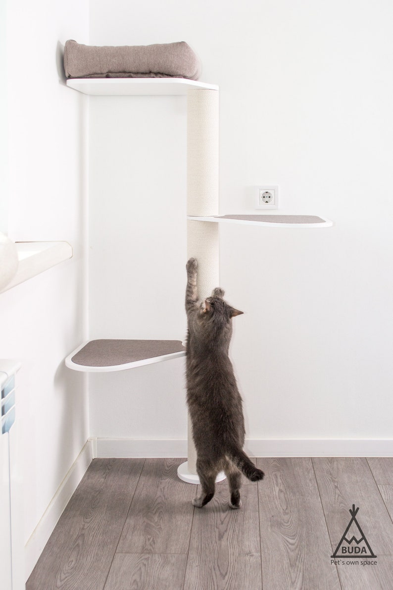 May include: A gray cat scratching a tall white cat tree with multiple platforms. The cat tree is in a corner of a room with white walls and a wood floor. The cat tree has a beige cushion on the top platform.