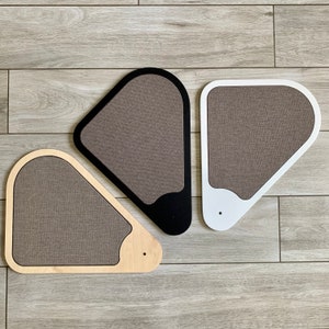 May include: Three cat scratching posts shaped like triangles. The posts are made of wood and covered in a brown fabric. The posts are in different colours: black, white, and natural wood.