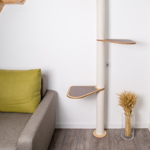 May include: A white cat tree with three platforms and a scratching post. The platforms are made of wood and have a gray fabric covering. The scratching post is covered in sisal rope. The cat tree is standing in a room with a gray couch and a vase of wheat.