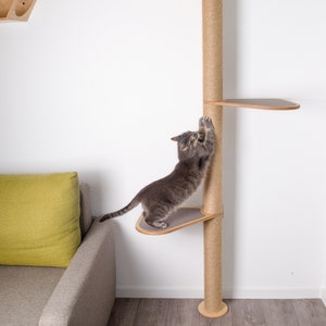 May include: A gray cat scratching a tall, sisal-wrapped scratching post with multiple platforms. The scratching post is made of wood and has a natural finish. The cat is standing on one of the platforms, which is made of wood and has a gray fabric covering. The scratching post is in front of a white wall and a gray couch with a green pillow.