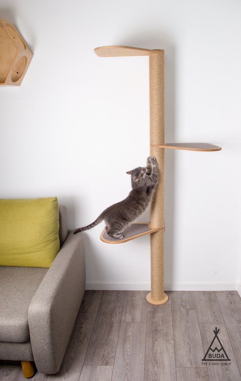 May include: A gray cat scratching a tall, sisal-wrapped scratching post with multiple platforms. The scratching post is made of wood and has a natural finish. The cat is standing on one of the platforms, which is made of wood and has a gray fabric covering. The scratching post is in front of a white wall and a gray couch with a green pillow.
