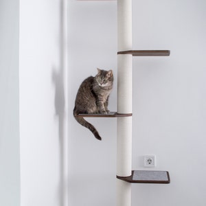 May include: A multi-level cat tree with a gray tabby cat sitting on a dark brown platform. The cat tree has a white, textured central post with several platforms and a gray carpeted base. The brand logo is visible at the bottom.