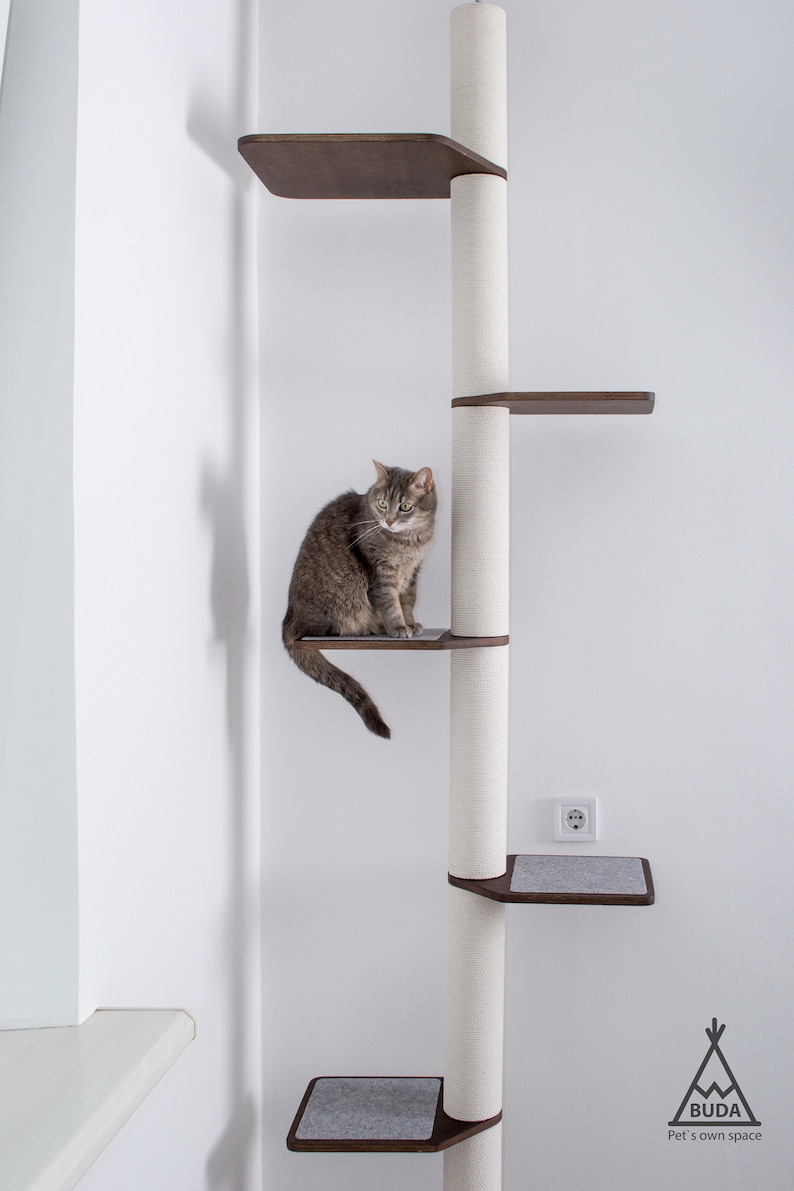 May include: A multi-level cat tree with a gray tabby cat sitting on a dark brown platform. The cat tree has a white, textured central post with several platforms and a gray carpeted base. The brand logo is visible at the bottom.