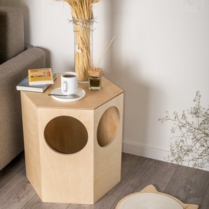 May include: A light wood hexagonal cat house with three round openings. The house is on a wood floor with a white cat scratching pad with a rope surface and a string attached. The text "BUDa Pet & own space" is on the scratching pad.
