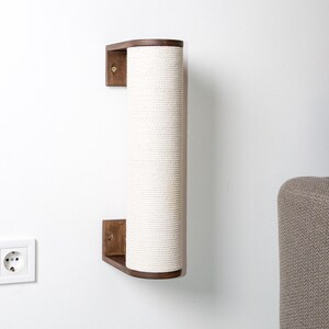 Pode incluir: Um arranhador de parede para gatos com uma cobertura de corda de sisal branca. O poste &eacute; de madeira e tem uma forma curva. Est&aacute; montado numa parede branca.