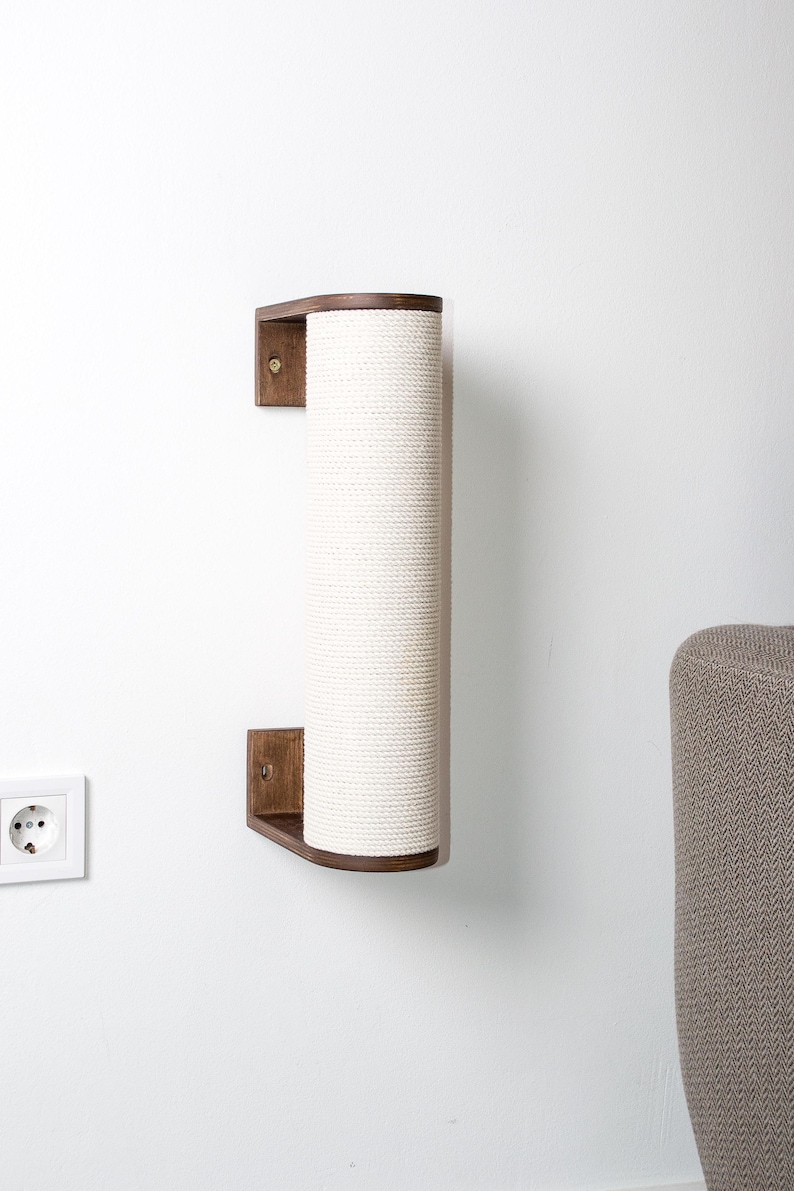 Pode incluir: Um arranhador de parede para gatos com uma cobertura de corda de sisal branca. O poste &eacute; de madeira e tem uma forma curva. Est&aacute; montado numa parede branca.