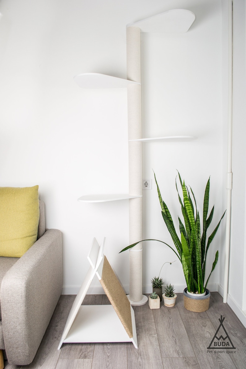 Puede incluir: &Aacute;rbol para gatos blanco con m&uacute;ltiples plataformas y un poste para rascar. El &aacute;rbol para gatos est&aacute; en una sala de estar con un sof&aacute; y una planta.