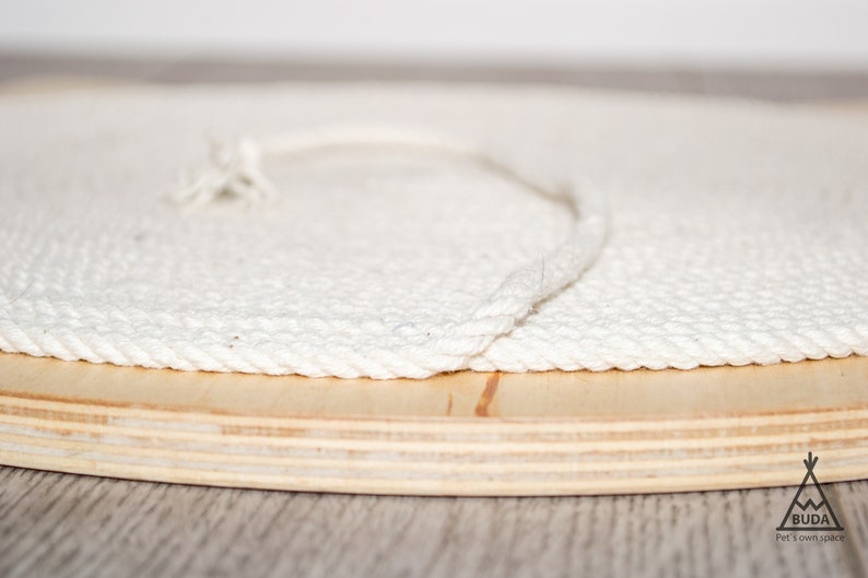 May include: Close-up of a white rope wrapped around the edge of a wooden round cat tree base. The base is made of plywood and has a natural wood finish.