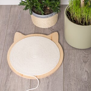 May include: A white rope cat scratching post with a wooden base shaped like a cat's head. The post is on a light gray wood floor.
