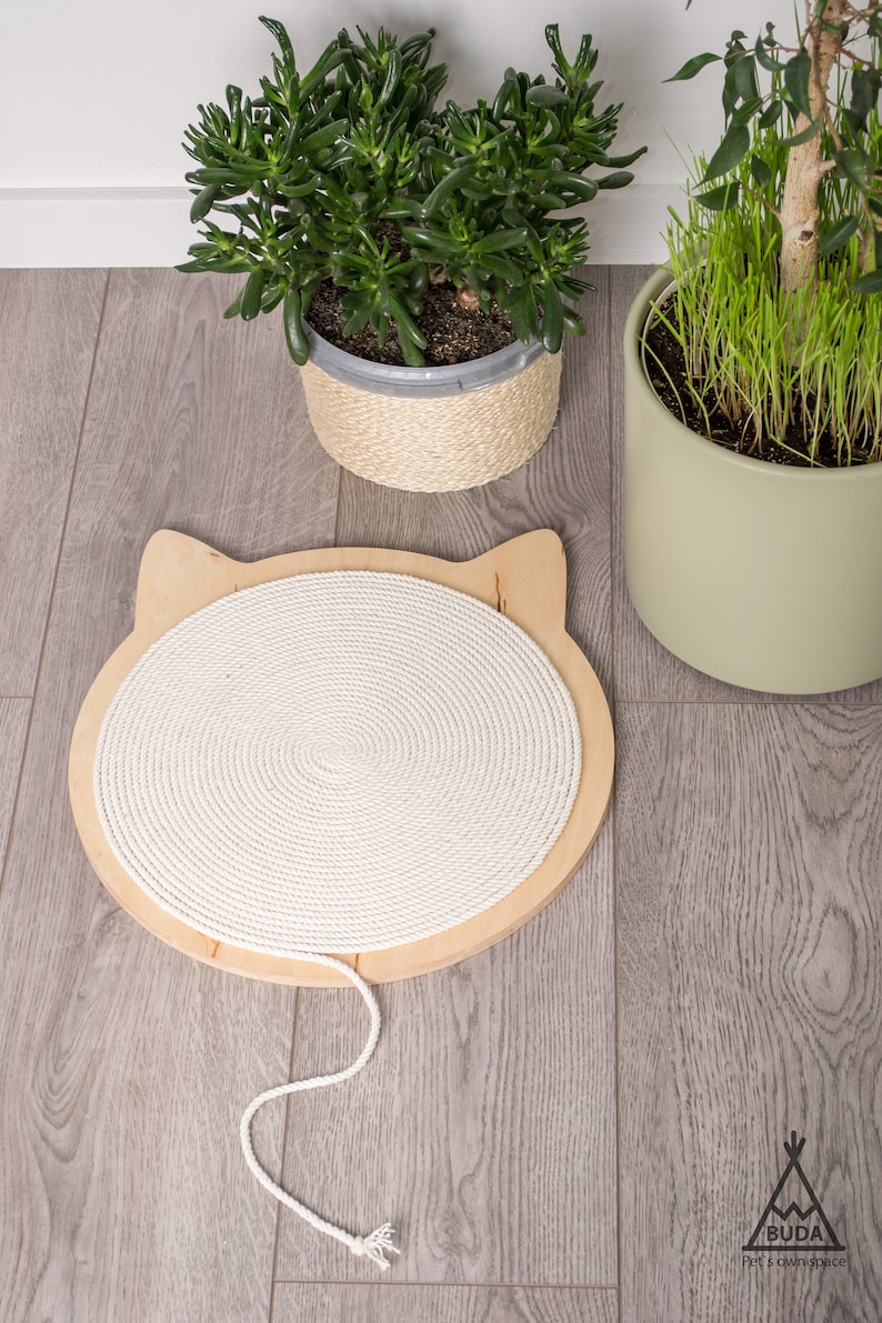 May include: A white rope cat scratching post with a wooden base shaped like a cat's head. The post is on a light gray wood floor.