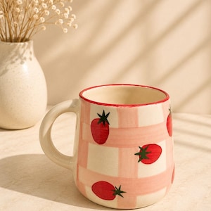 May include: A ceramic mug featuring a hand-painted light pink checkered pattern across the exterior. The mug is decorated with small red strawberry illustrations, each topped with a green stem. The rim is accented with a thin red painted line.
