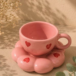 Może przedstawiać: Różowy ceramiczny zestaw filiżanki ze spodkiem. Filiżanka posiada zaokrąglone ucho i małe motywy czerwonych serc. Spodek ma falisty, kwiatowy brzeg z pasującymi detalami czerwonych serc umieszczonymi na płatkach.
