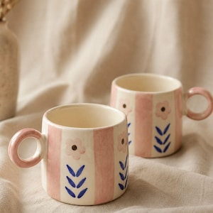 May include: Two ceramic mugs featuring a cream-coloured base with vertical pink stripes. Each mug is decorated with hand-painted pink flowers with dark centres and blue leaf motifs. The mugs have a rounded pink handle and a cylindrical shape.