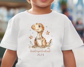 Könnte beinhalten: Weißes T-Shirt mit einer braunen Dinosaurier-Illustration und dem Text "kindergartenkind 2024". Ein kleines Kind trägt das Shirt.