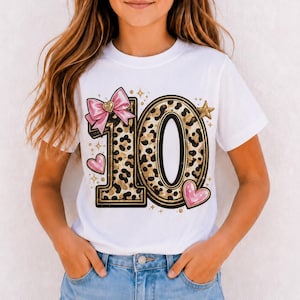 Könnte beinhalten: Ein weißes Kurzarm-T-Shirt mit einer großen Grafik der Zahl 10 in Leopardenmuster. Das Design enthält eine rosa Schleife, rosa Herzen und goldene Sterne. Das T-Shirt wird mit hellblauen Jeansshorts und einer schwarzen Sonnenbrille kombiniert.