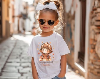 Pode incluir: Uma camiseta branca com uma ilustração em aquarela de uma criança vestida de bombeiro. O texto "kindergartenkind Charlotte 2025" está impresso na camiseta.