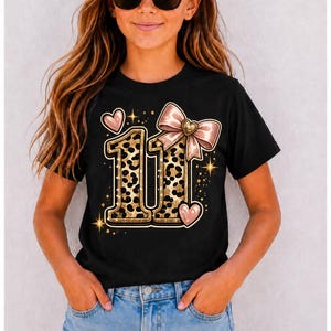 Könnte beinhalten: Ein schwarzes Kurzarm-T-Shirt mit einer großen Grafik der Zahl 11 in einem gold-schwarzen Leopardenmuster. Das Design ist auf der Vorderseite mit einer hellrosa Schleife, zwei kleinen rosa Herzen und goldenen Sternenfunken verziert.