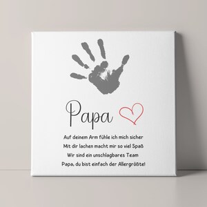 May include: A white canvas print featuring a gray handprint above the word "Papa" in elegant script. A red heart is drawn next to the word. The print includes German text expressing love and appreciation.