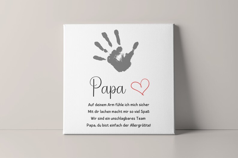 May include: A white canvas print featuring a gray handprint above the word "Papa" in elegant script. A red heart is drawn next to the word. The print includes German text expressing love and appreciation.