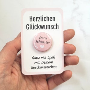 May include: A pink card with German text and a pink button. The button says "Große Schwester" and has a heart design. The card is held in a hand. The card's text translates to "Congratulations" and "Have fun with your siblings".