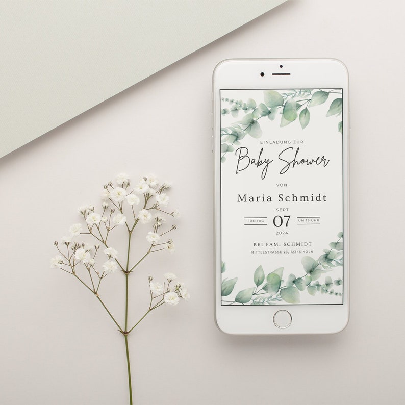 May include: A white smartphone displays a baby shower invitation with eucalyptus leaf accents. The invitation text reads "Baby Shower" and includes details for Maria Schmidt's event on September 7, 2024. A sprig of white flowers is in the foreground.