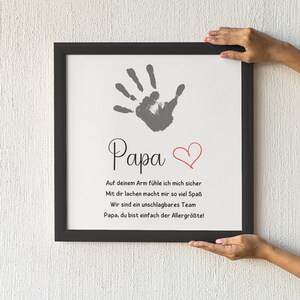 May include: A framed print with a handprint and the word "Papa" in script. A red heart is next to the word. The print includes a message in German. The frame is black.