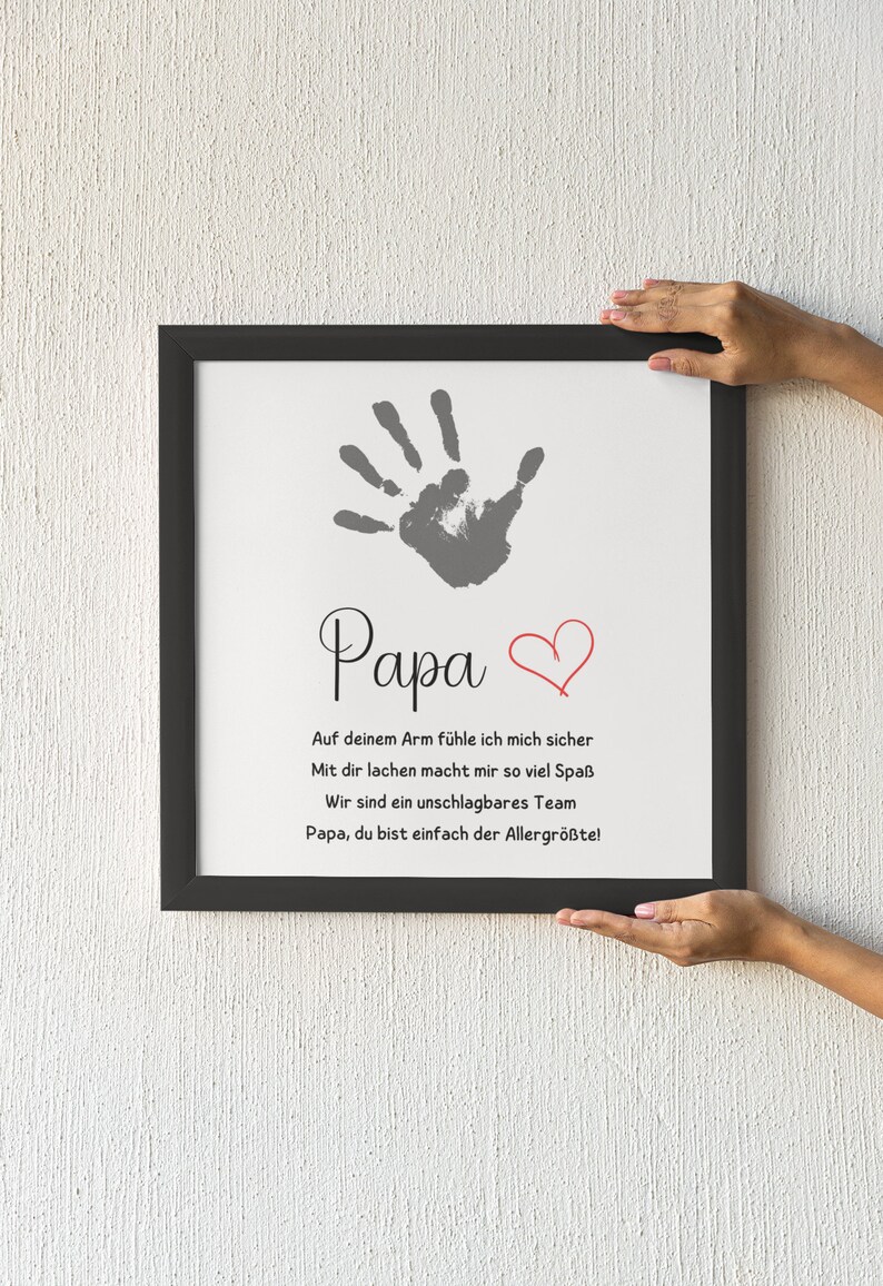 May include: A framed print with a handprint and the word "Papa" in script. A red heart is next to the word. The print includes a message in German. The frame is black.