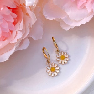 Peut inclure: Une paire de boucles d'oreilles cr&eacute;oles dor&eacute;es avec un charme de marguerite blanche avec un centre jaune. Les boucles d'oreilles sont sur une assiette blanche avec des fleurs roses en arri&egrave;re-plan.