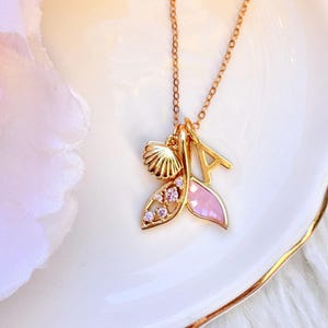 May include: A gold-coloured necklace featuring a seashell charm, a pink enamel charm, and the letter "A" charm. The pink charm is shaped like a mermaid's tail and is decorated with small pink crystals. The necklace is on a white plate.