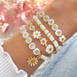 May include: Four white beaded bracelets with gold lettering spelling out "DREAMER" and "MAMA". Each bracelet has small white daisy charms with yellow centres.