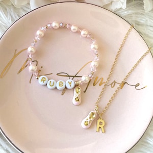 May include: A pink and gold necklace and bracelet set with a ballerina shoe charm and the name "Ruby" spelled out in white beads.
