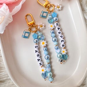 May include: Two personalized keychains with gold-colored hardware. Each keychain features a beaded strand with the name "SARAH" and "HARPER" spelled out in white letter beads. The strands include blue beads, daisy accents, and small book charms.