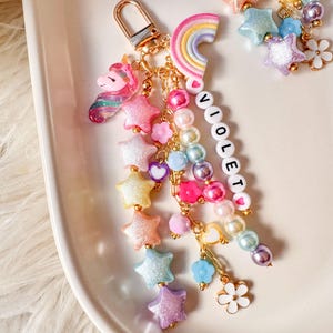 May include: A gold-toned keychain featuring a unicorn charm, a rainbow, and multiple strands of colorful beads. The beads include glittery stars, round pearls, small flowers, and white letter beads spelling VIOLETO, all attached to a gold lobster clasp.