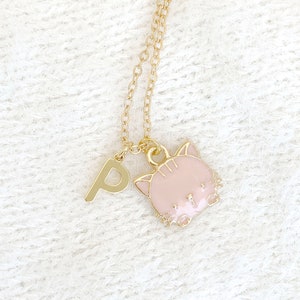 May include: A gold-toned metal necklace featuring a delicate chain with two charms: a capital letter P and a light pink enamel cat head with gold-toned outlines. The cat charm displays simple facial features including ears, whiskers, and eyes.