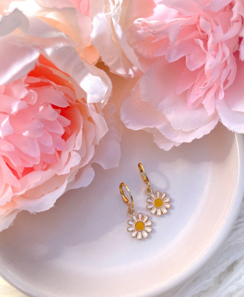 Peut inclure: Une paire de boucles d'oreilles cr&eacute;oles dor&eacute;es avec un charme de marguerite blanche avec un centre jaune. Les boucles d'oreilles sont sur une assiette blanche avec des fleurs roses en arri&egrave;re-plan.