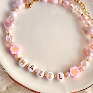May include: A pink beaded necklace spelling "HAZEL" in white letter beads with gold lettering. The necklace features pink heart, star, and flower-shaped beads, along with clear and pearl beads. A gold chain and clasp complete the design.