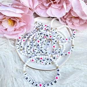 May include: A stack of white beaded bracelets with black lettering and pink hearts spelling out "IBIZA CREW 24" and other variations. The bracelets are on a white surface.