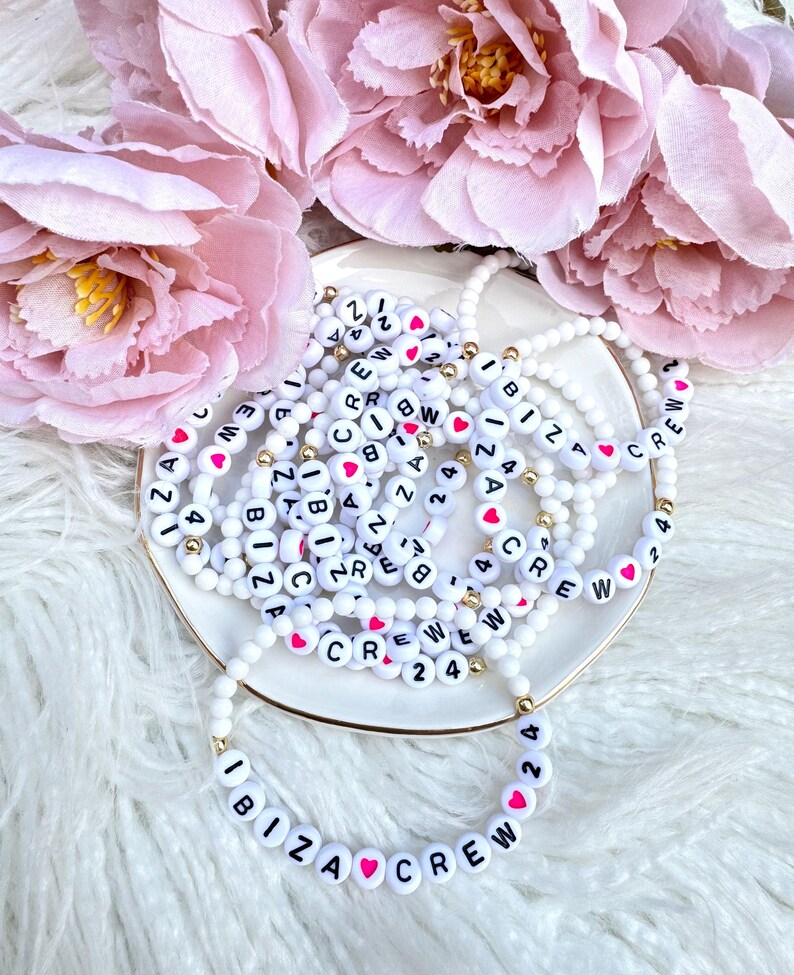 May include: A stack of white beaded bracelets with black lettering and pink hearts spelling out "IBIZA CREW 24" and other variations. The bracelets are on a white surface.