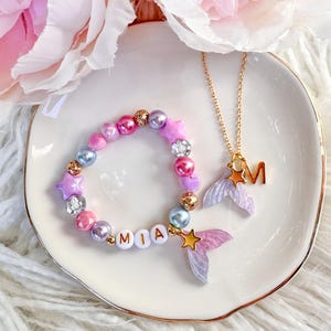 May include: A beaded bracelet featuring pink, purple, and blue beads, star-shaped charms, and white letter beads spelling MIA, finished with a mermaid tail charm. A gold-tone chain necklace with a letter M charm and a mermaid tail pendant.