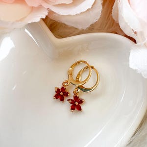 May include: Gold-coloured hoop earrings with dangling red flower charms. The earrings are displayed on a white, heart-shaped dish, with soft pink and white flowers in the background.
